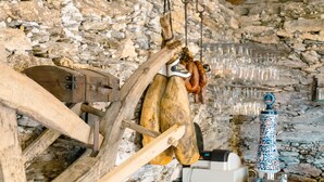 A rustic kitchen with a stone wall, a wooden meat curing rack, hanging sausages, and a decorative ceramic pot. - Hotel Atalaya II (Sanxenxo)