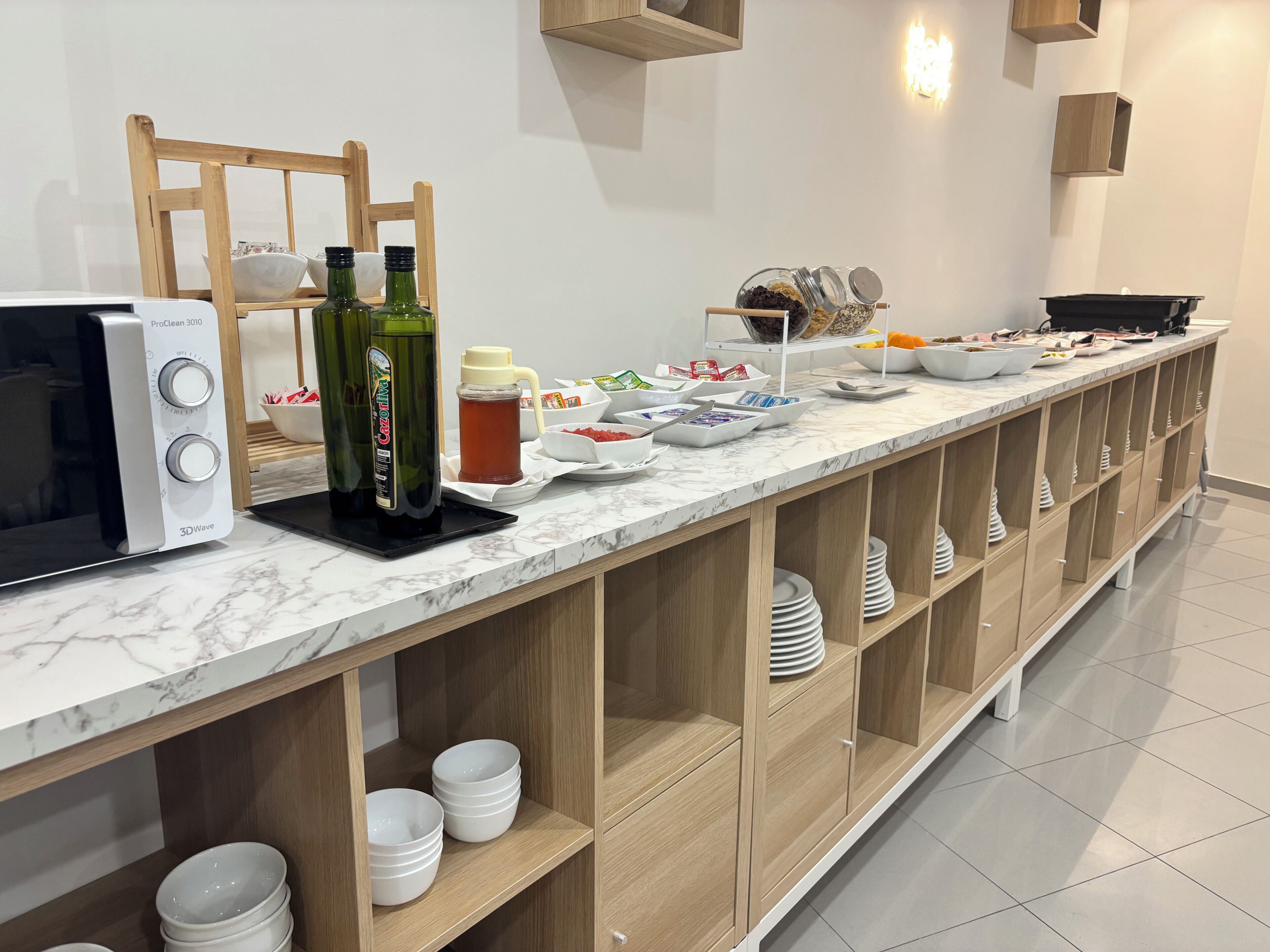 A breakfast buffet setup with a ProClean 3010 microwave, green bottles, a honey dispenser, and various food items on a marble counter.