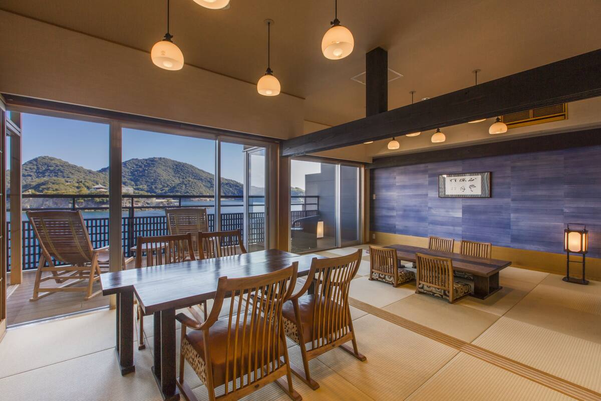 A spacious room with a large dining table and chairs, a small seating area, and a view of the mountains and water through large windows.