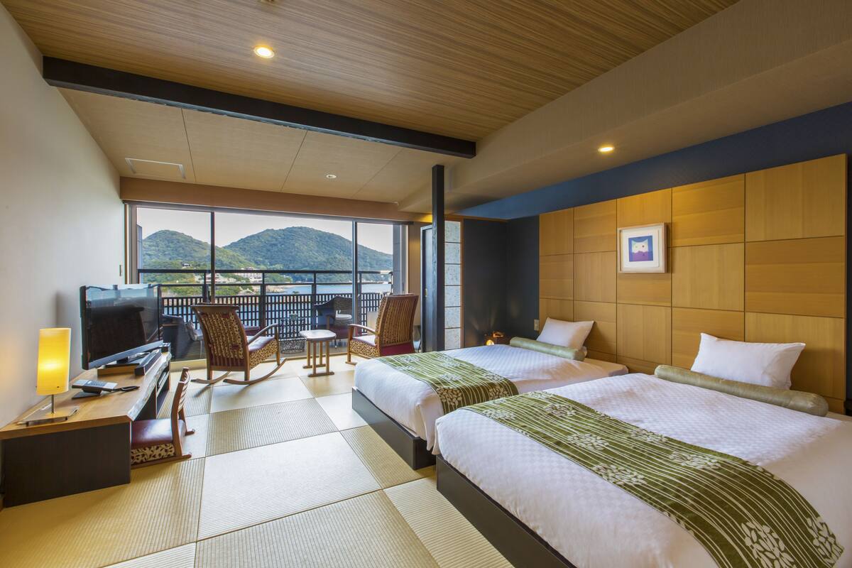 A hotel room with two beds, a TV, a desk, and a balcony with a view of mountains.