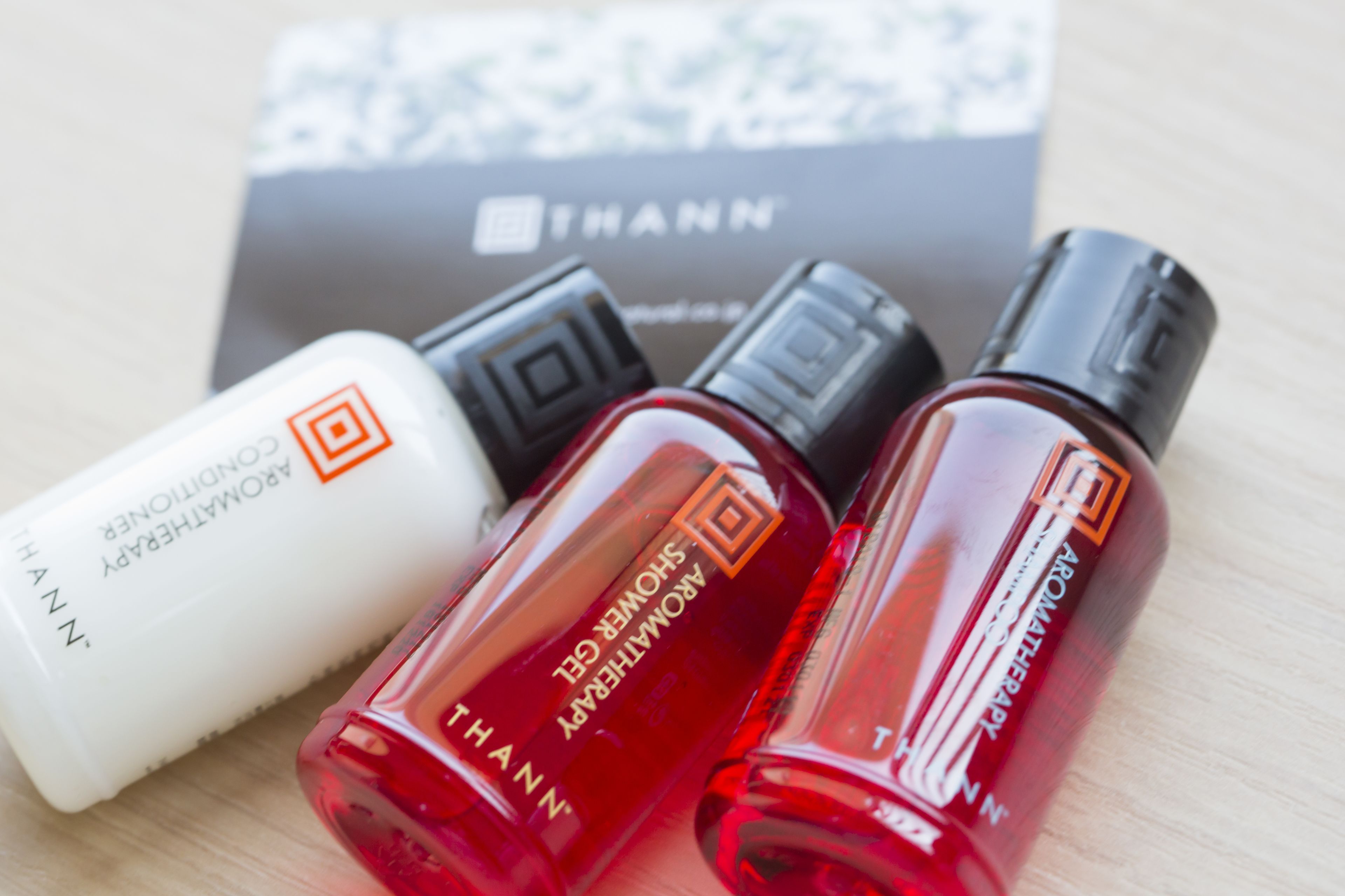 Three perfume bottles with labels, one white and two red, placed on a wooden surface with a THANN brand box in the background.