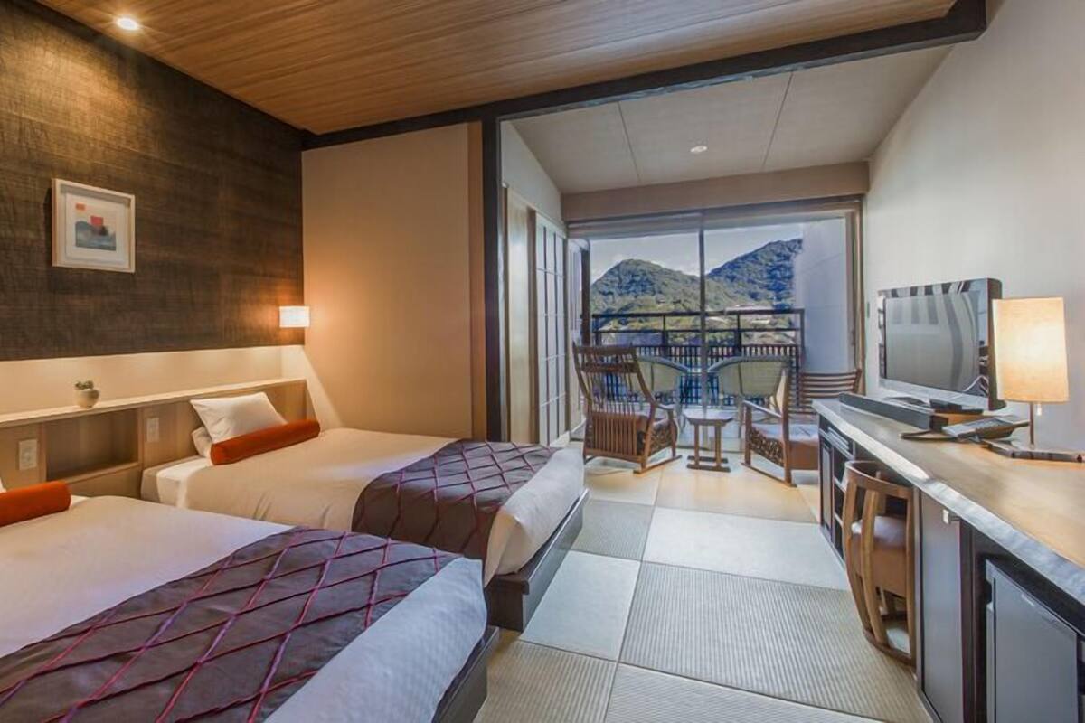 A hotel room with two beds, a desk with a TV, and a balcony with a mountain view.