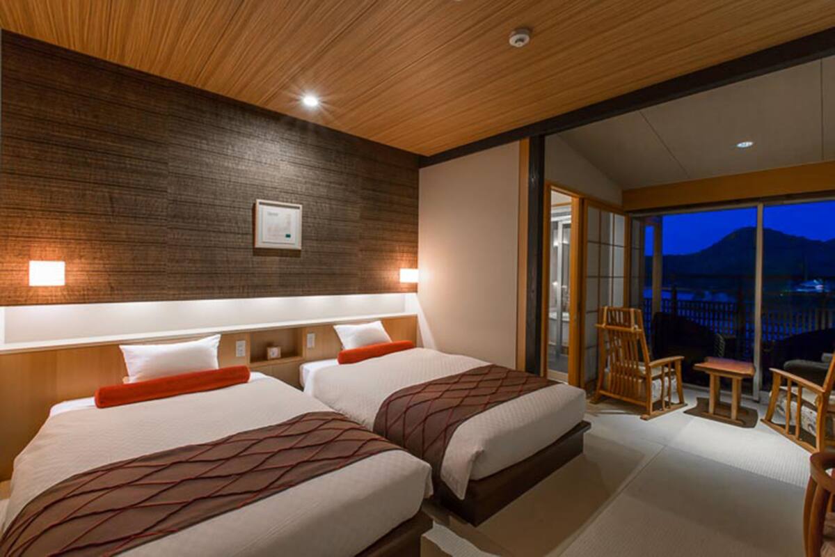 A hotel room with two beds, a wooden ceiling, a night view balcony, and a sliding door leading to an outdoor area.