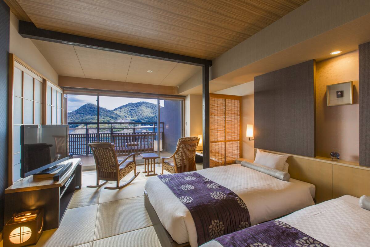 A hotel room with two beds, a TV, a desk, and a balcony with a view of mountains.