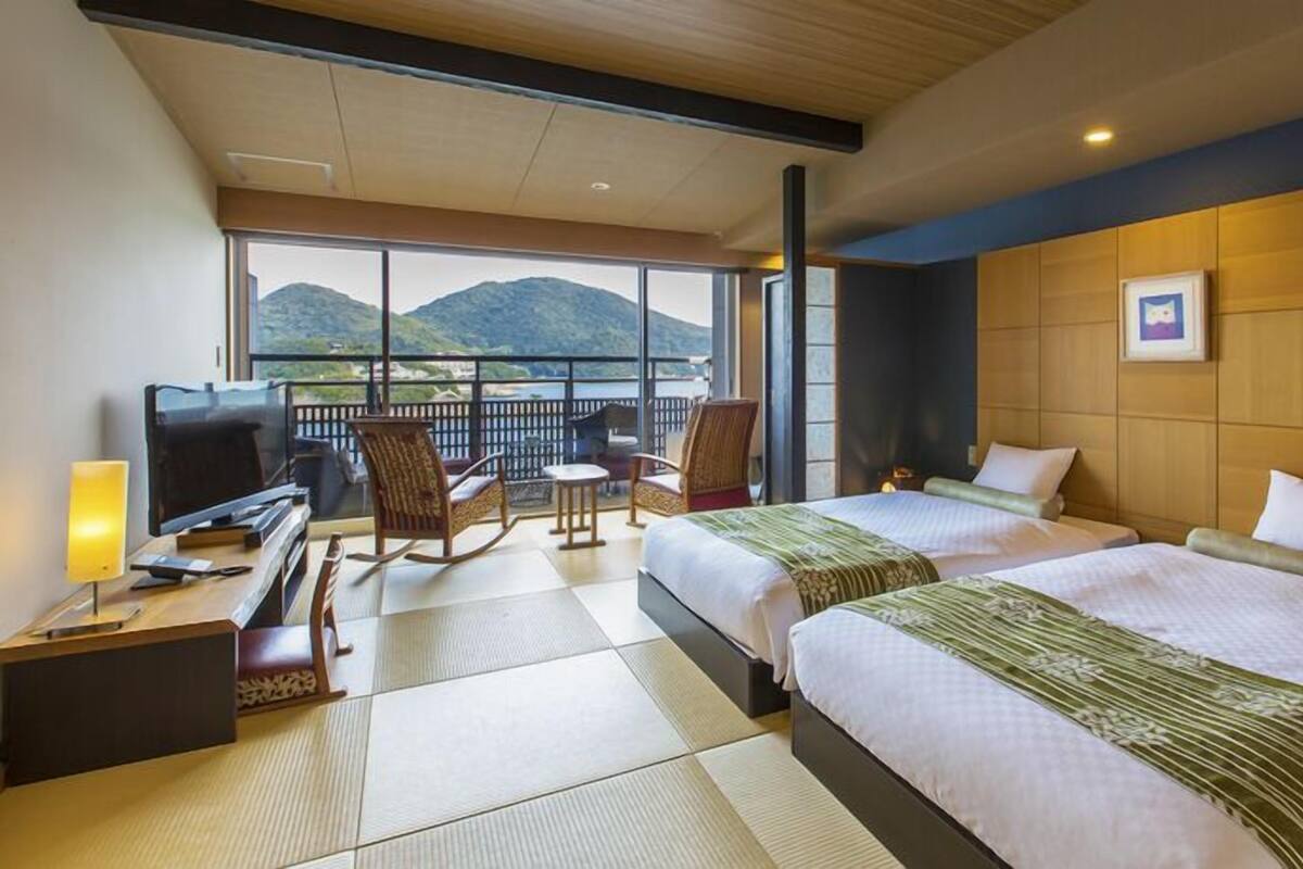 A hotel room with two beds, a TV, a desk, a chair, and a balcony with a mountain view.