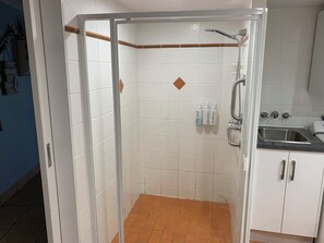 A shower with a glass door, white tiled walls, and a tiled floor. There are toiletries on the wall and a sink with a faucet. - Northpoint Holiday Apartments (Alexandra Headland)