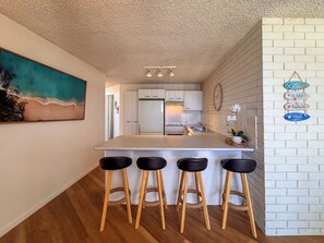 A kitchen with a white island, bar stools, a refrigerator, and a wall-mounted clock. - Northpoint Holiday Apartments (Alexandra Headland)