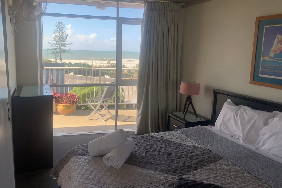 A hotel room with a bed, a nightstand, a lamp, a window with a view of the beach, and a fan.