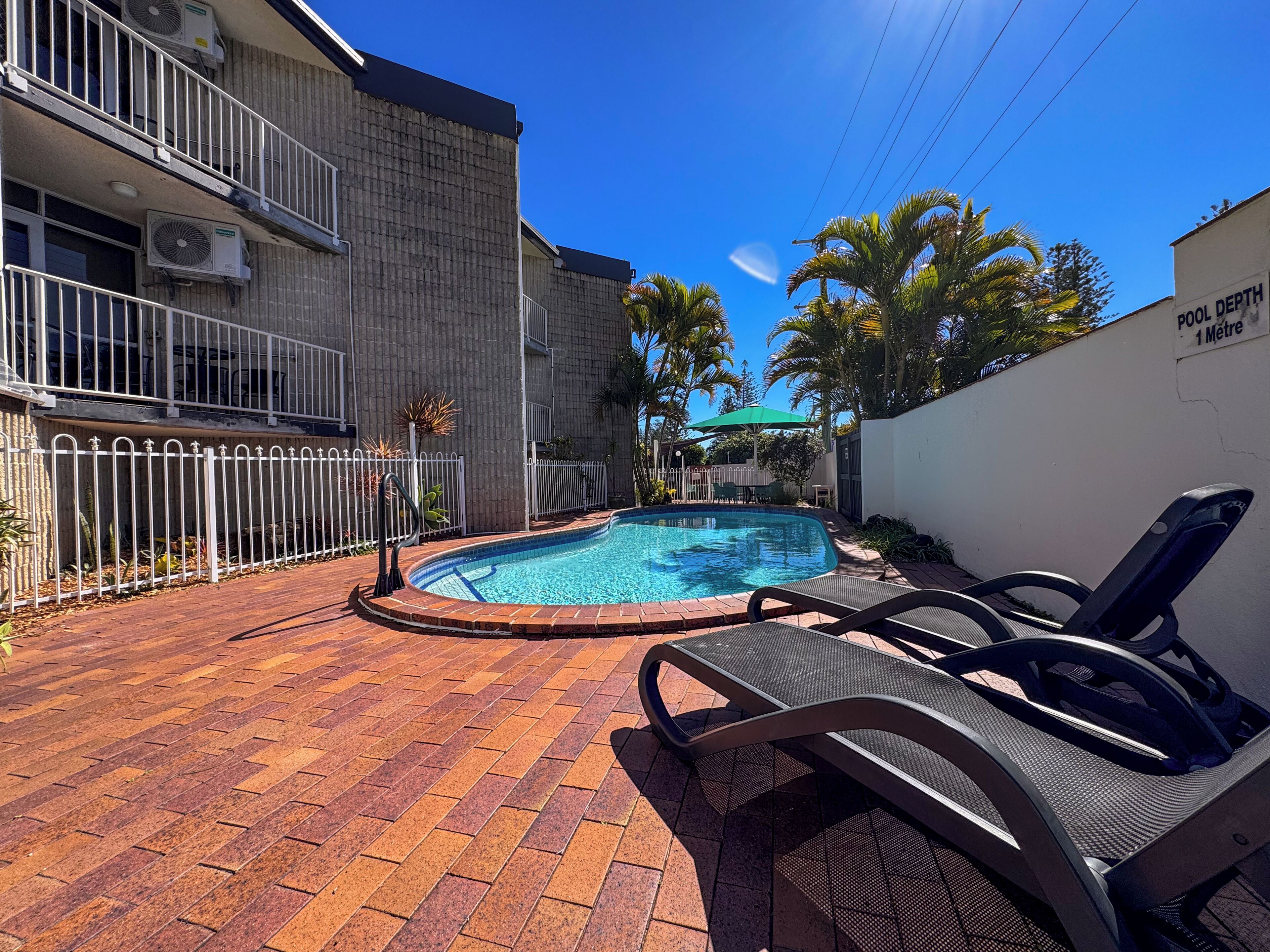 Une piscine avec une terrasse en briques, entourée d’une clôture blanche, et un bâtiment doté de balcons et de climatiseurs.