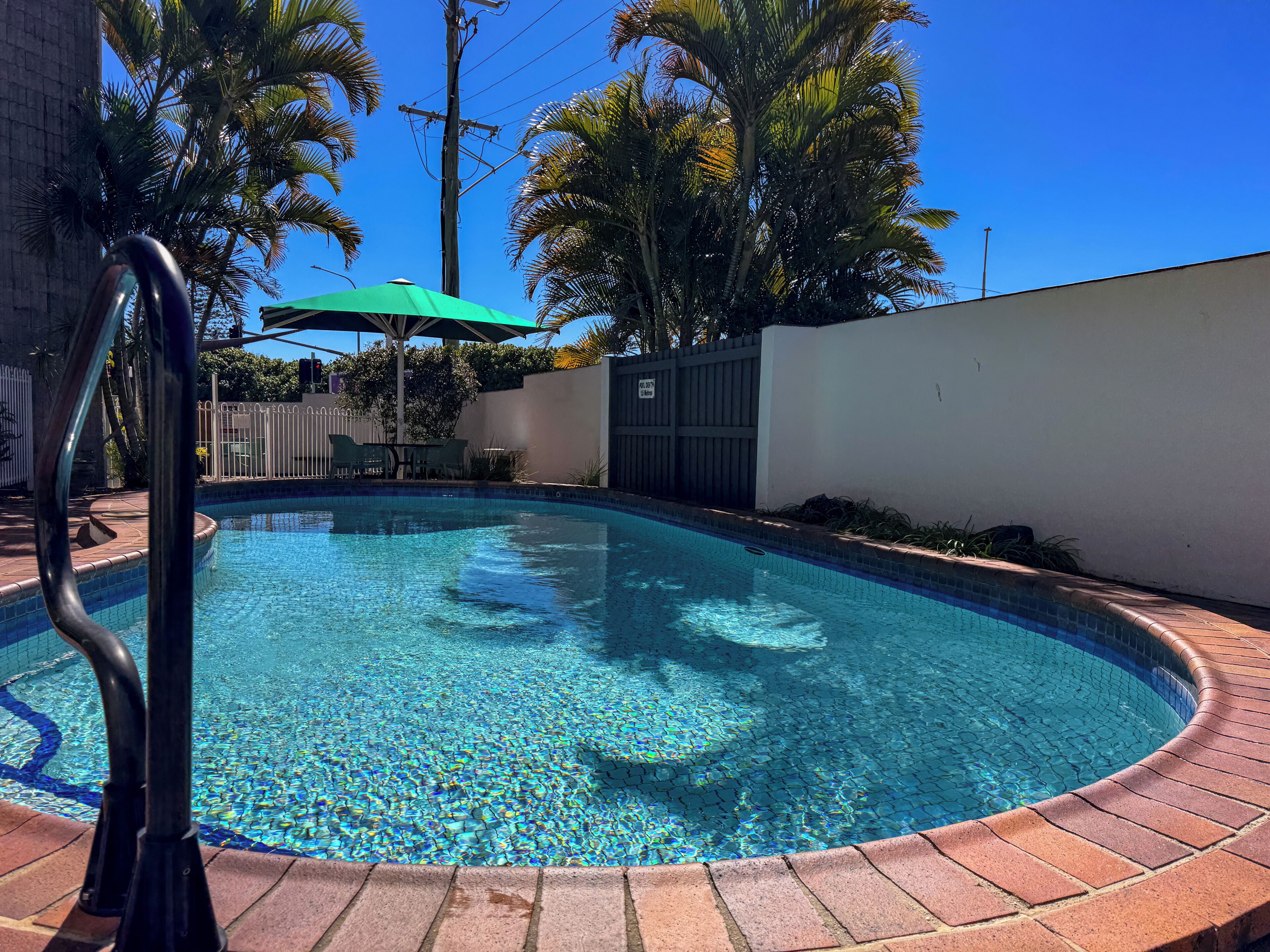 Una piscina con borde de azulejos, rodeada por un foso de ladrillo, palmeras y una pared blanca.