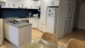 A modern kitchen with white cabinets, a stainless steel sink, and a built-in oven. There is a refrigerator with magnets and a small island with a glass top. - Northpoint Holiday Apartments (Alexandra Headland)