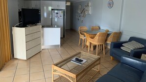 A modern living room with a blue sofa, a coffee table, wicker chairs, and a kitchen area with a refrigerator and microwave. - Northpoint Holiday Apartments (Alexandra Headland)
