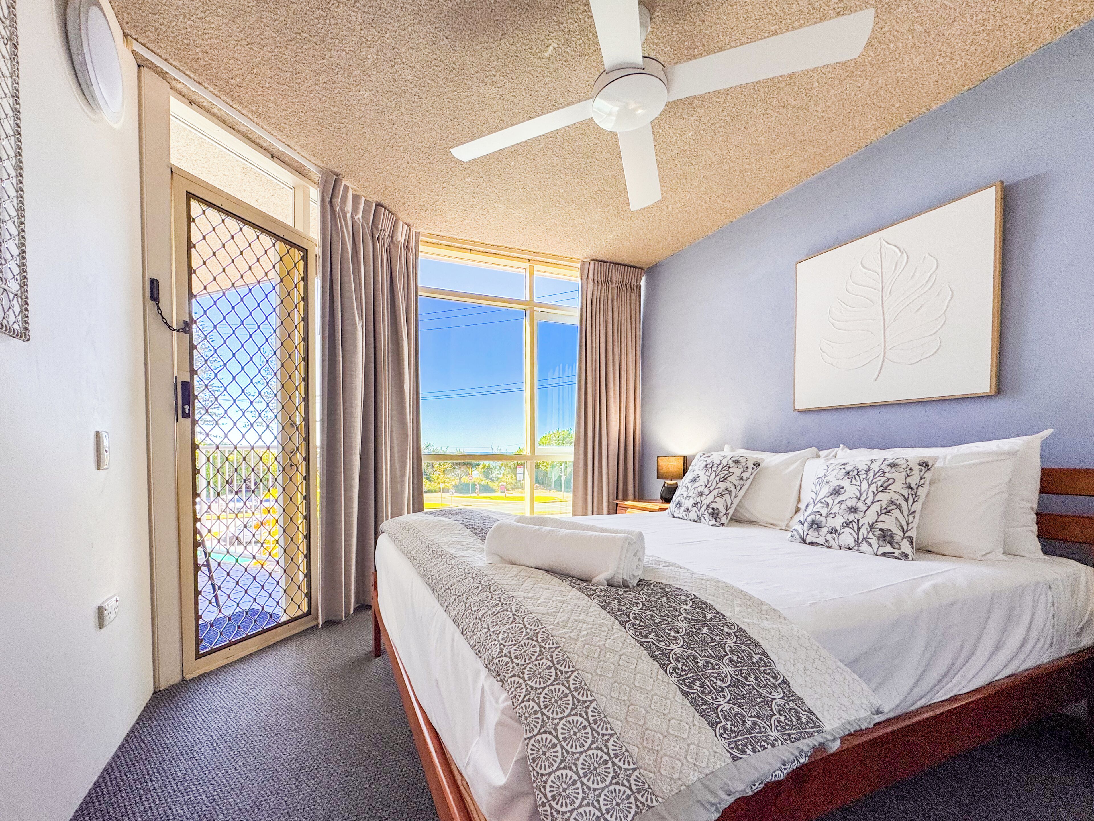 A hotel room with a bed, a ceiling fan, a window with curtains, and a door with a chain.