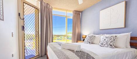 A hotel room with a bed, a ceiling fan, a window with curtains, and a door with a chain.