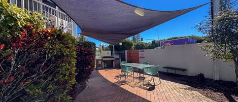 A patio area with a table and chairs, surrounded by a brick walkway and a shaded area.