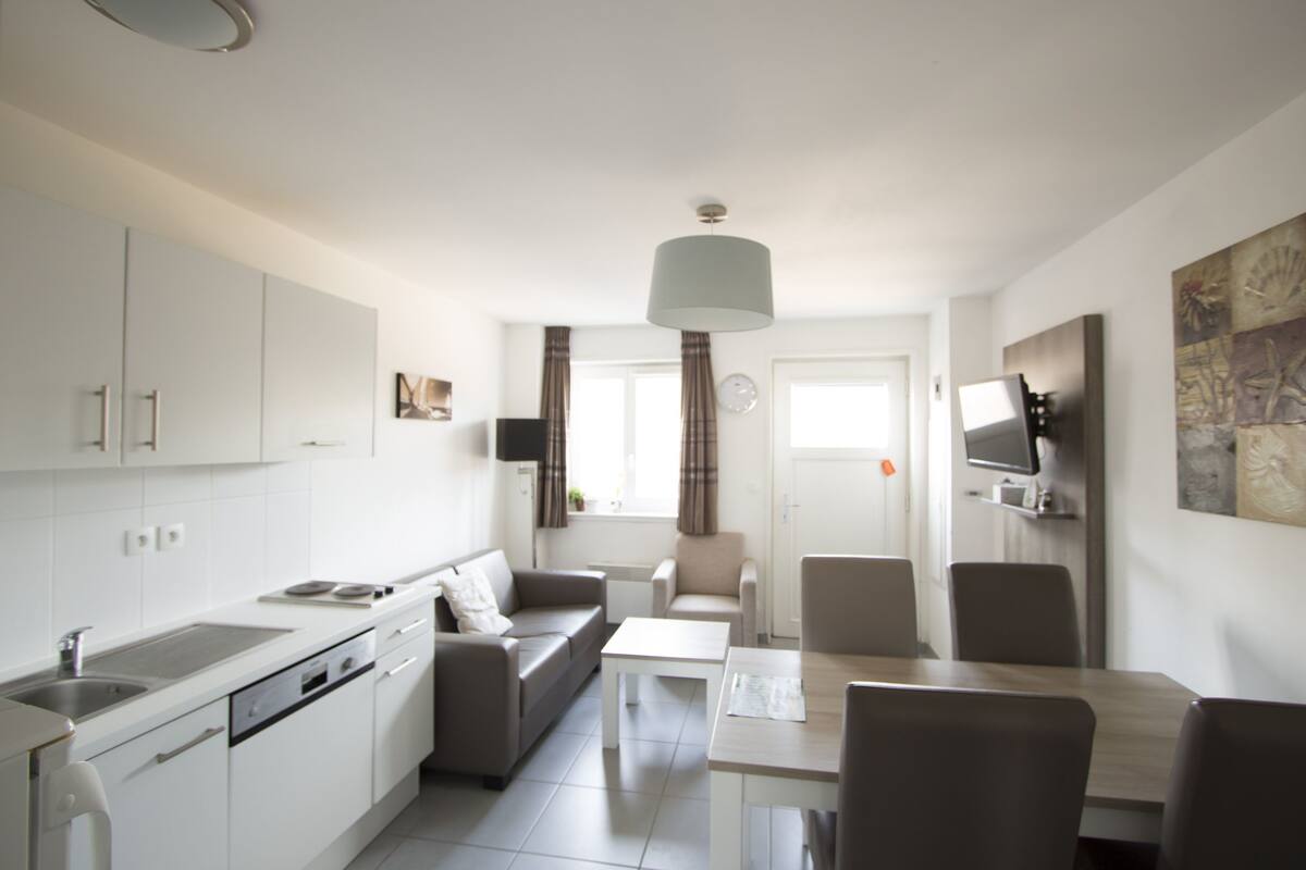 Une cuisine moderne avec des meubles blancs, un coin repas avec une table et des chaises, et un salon avec un canapé et un téléviseur.