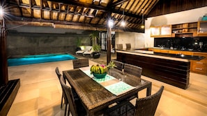 An outdoor dining area with wicker chairs and a table, a kitchen with wooden cabinets, and a swimming pool in the background. - Gili Villas (Gili Trawangan)