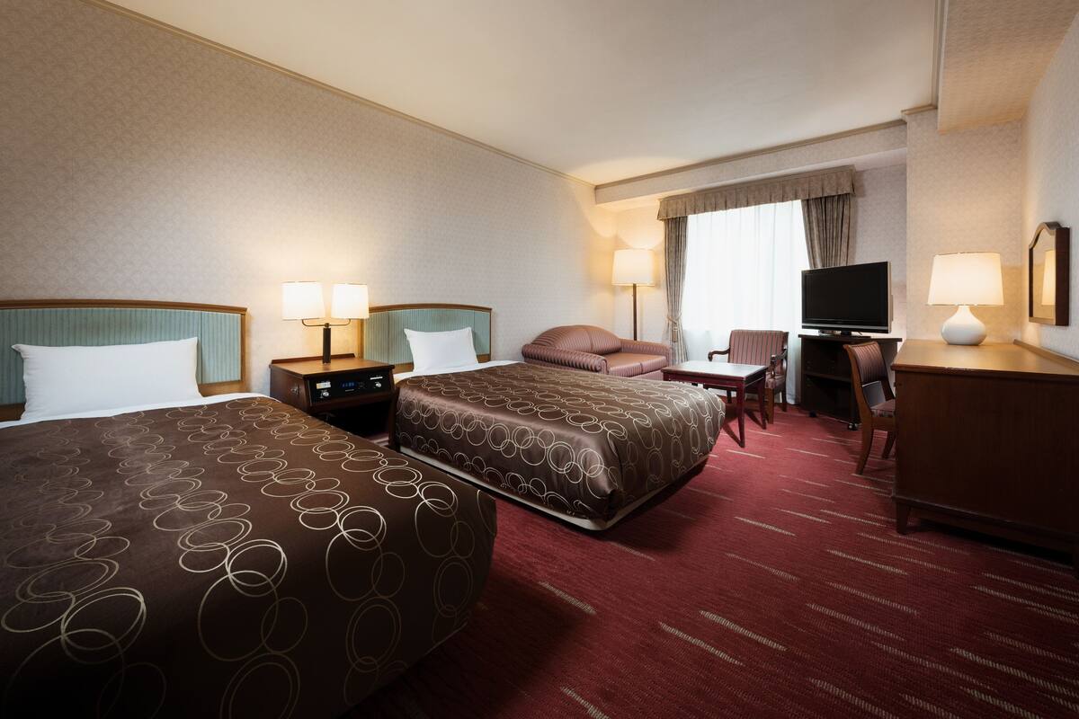 A hotel room with two beds, a desk, a television, and two lamps.
