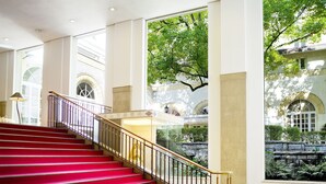 Eine imposante Treppe mit roten Teppichen führt zu einem Balkon mit Blick auf einen Garten und ein Gebäude mit Bogenfenstern.
