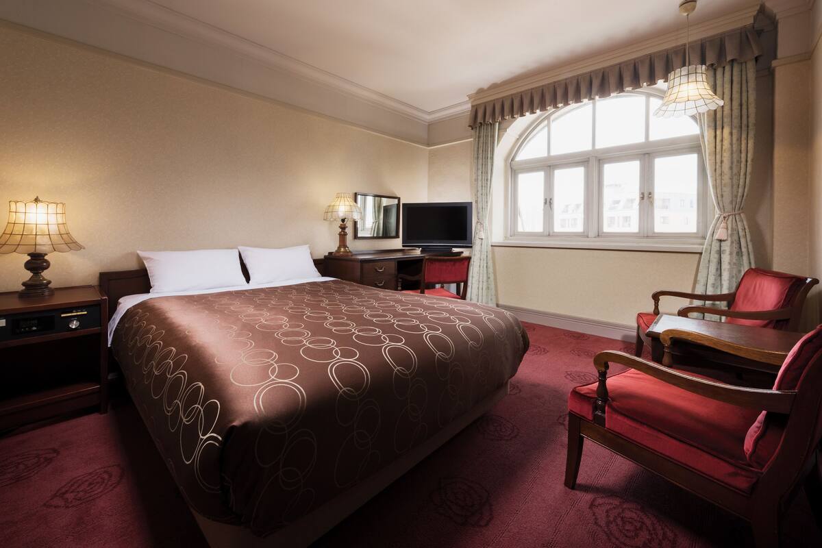 A hotel room with a large bed, a desk with a chair, a television, and a window with curtains.