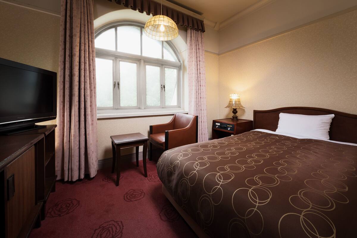A hotel room with a large bed, a television, a desk, and a window with curtains.
