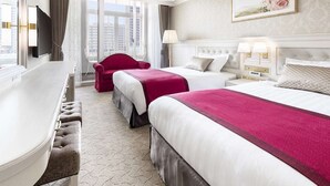 Ein Hotelzimmer mit zwei Betten, einer Sitzecke mit einem roten Sessel, einem Schreibtisch und einem großen Fenster mit Blick auf die Stadt.