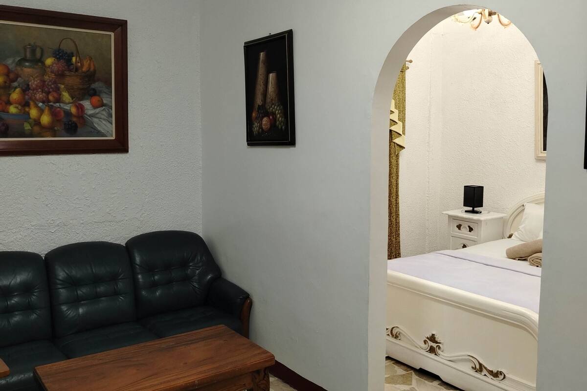 A room with a black leather sofa, a wooden coffee table, a painting of fruits, and a bed with a white headboard.