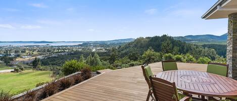 A wooden deck with a round table and chairs, overlooking a scenic landscape with hills and a body of water.