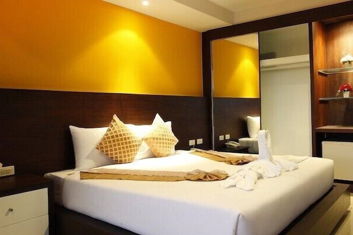 A hotel room with a large bed, a nightstand, a dresser, a mirror, and a shelf with a lamp.