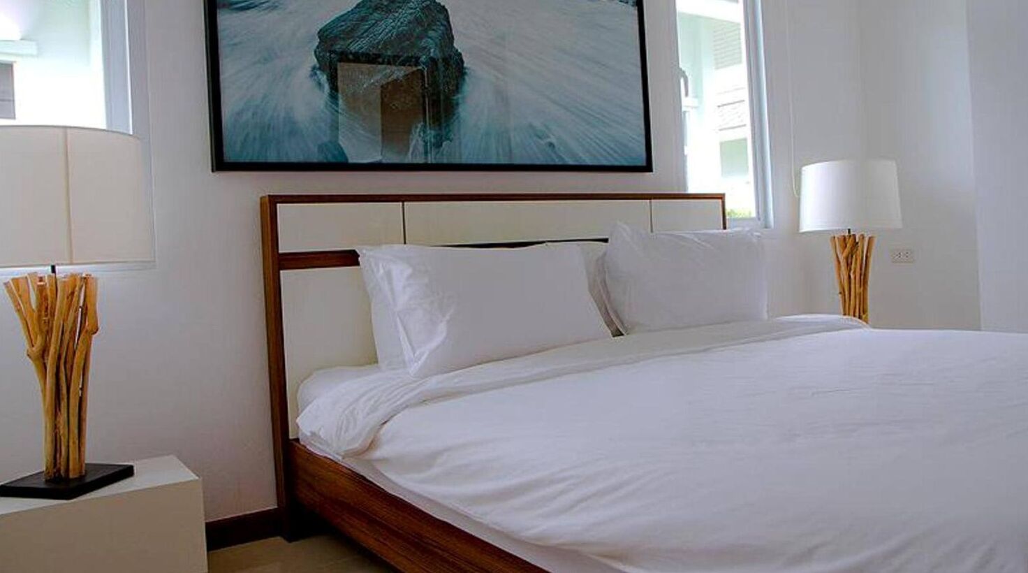 A neatly made bed with white linens, a wooden headboard, and a painting above it.