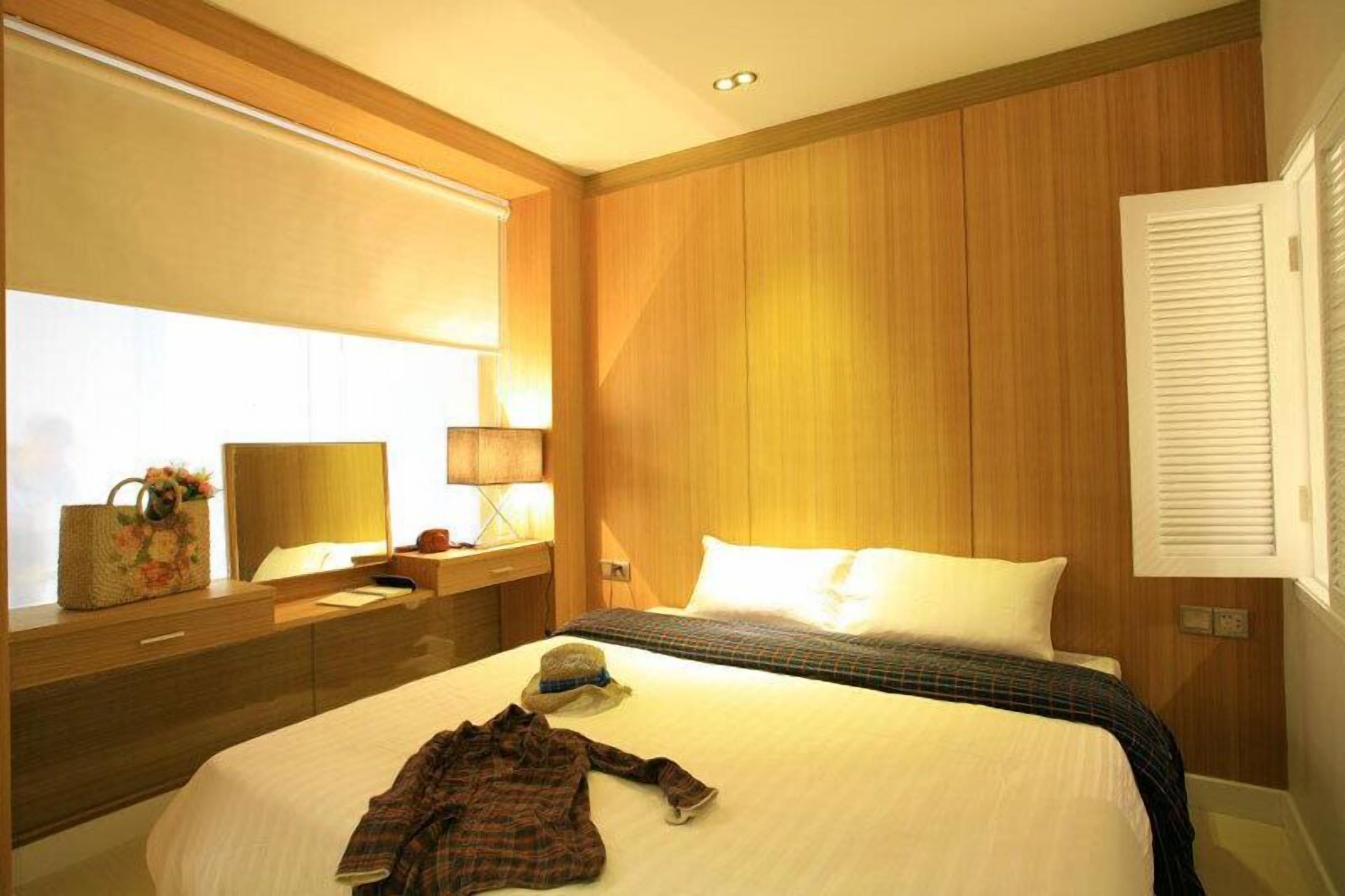 A hotel room with a large bed, wooden paneling, a nightstand with a lamp, and a window with blinds.