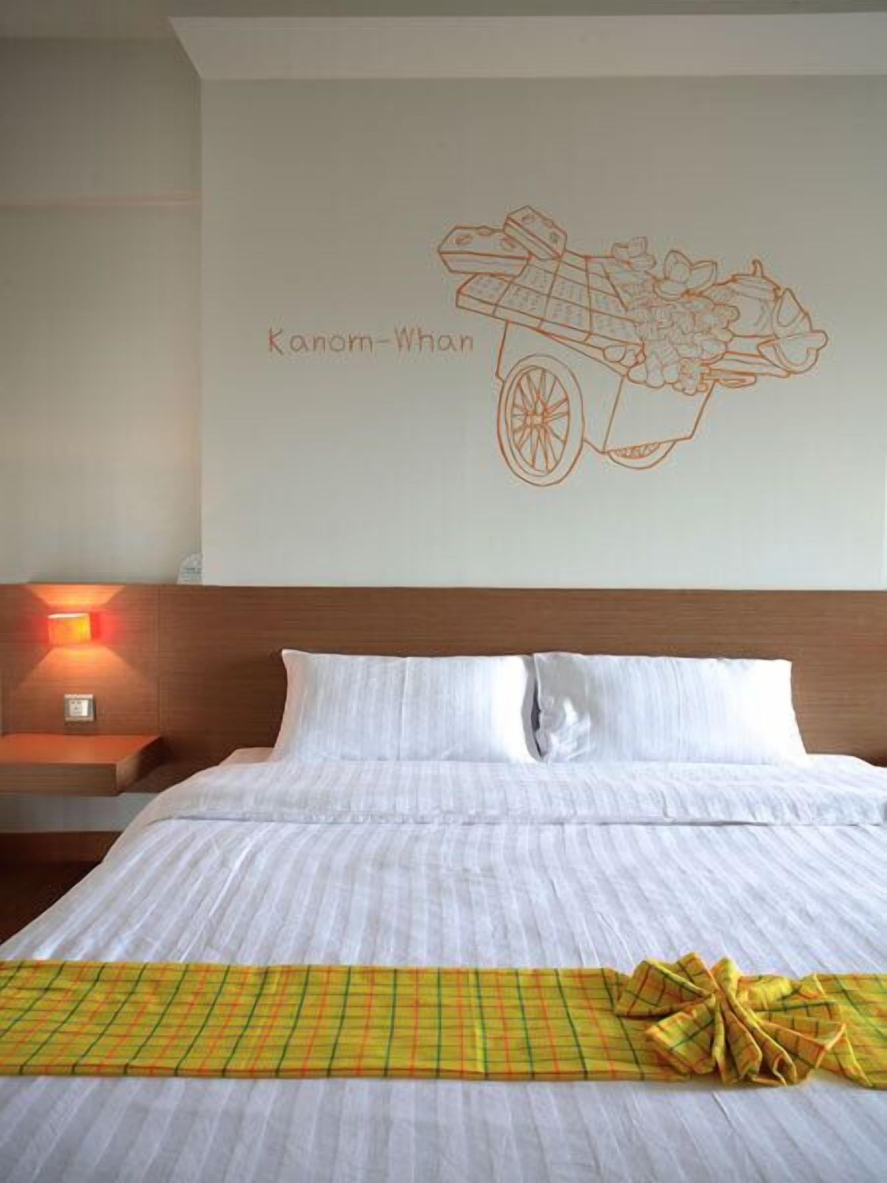 Habitación de hotel con cama, mesita de noche con lámpara y decoración de pared que muestra un carrito con el texto 'Kanom-Whan'.