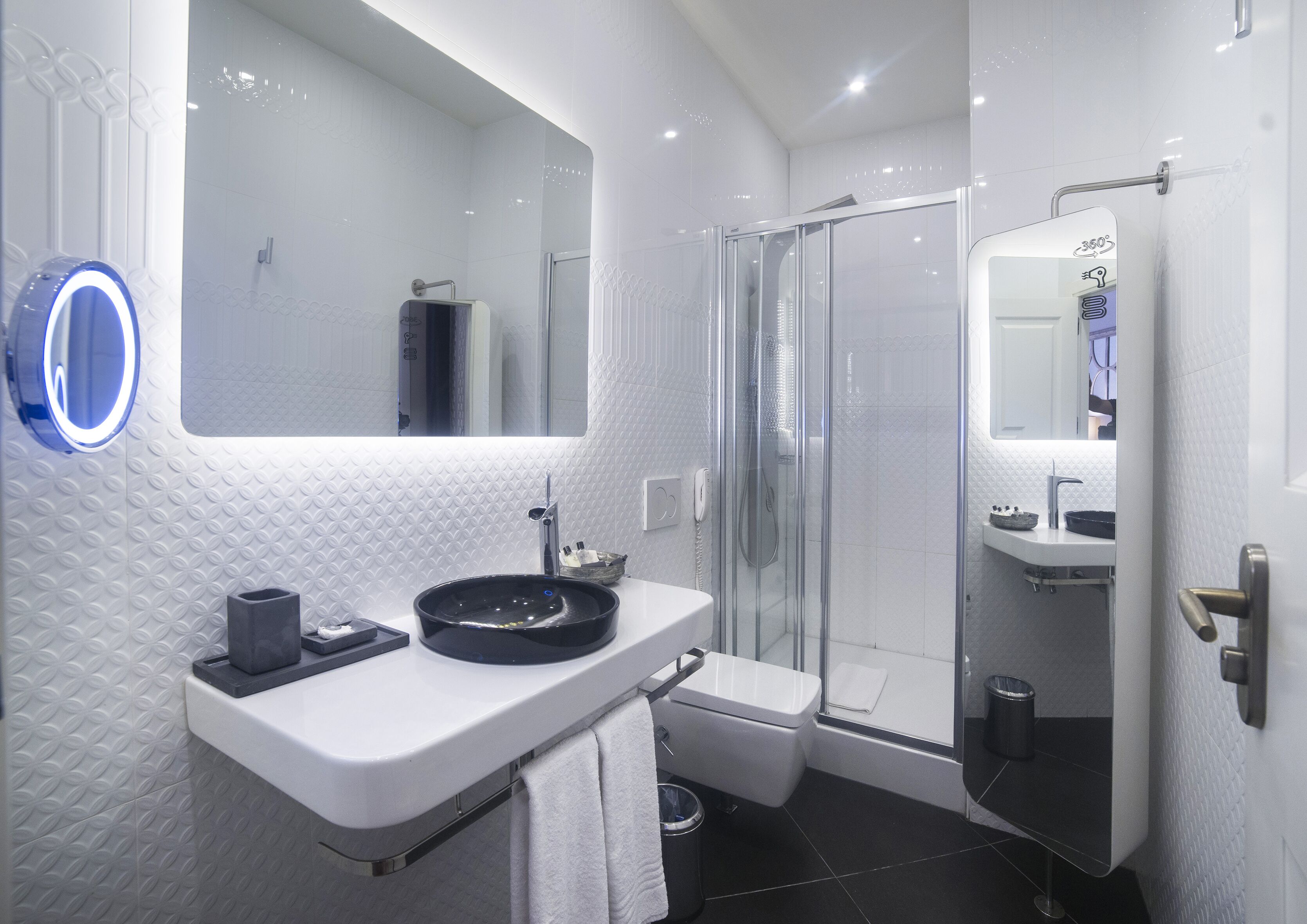 A modern bathroom with a glass-enclosed shower, a white sink with a black basin, and a large mirror with a circular light.