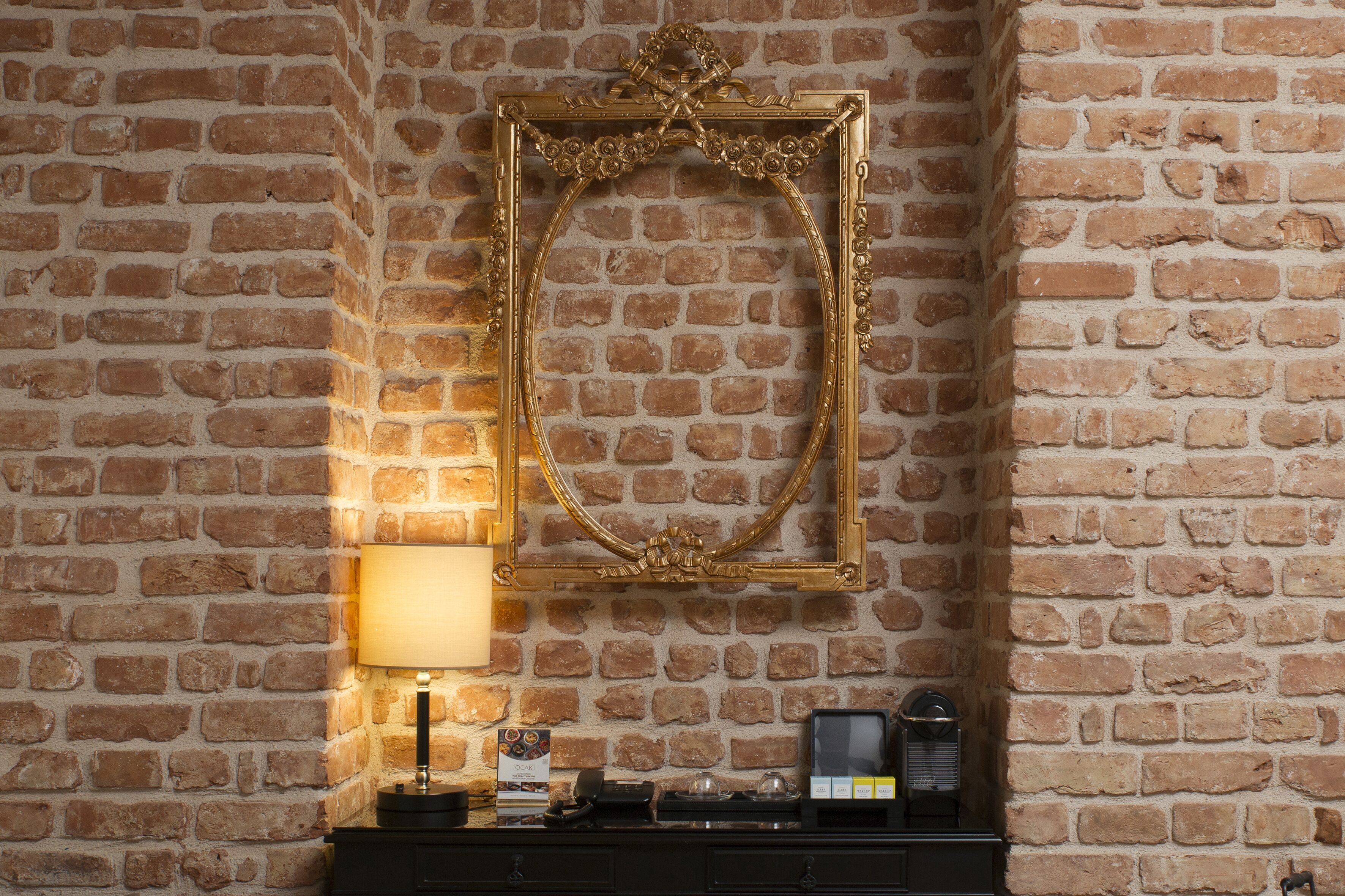 A brick wall with a gold-framed mirror and a small table with a lamp, books, and a radio.