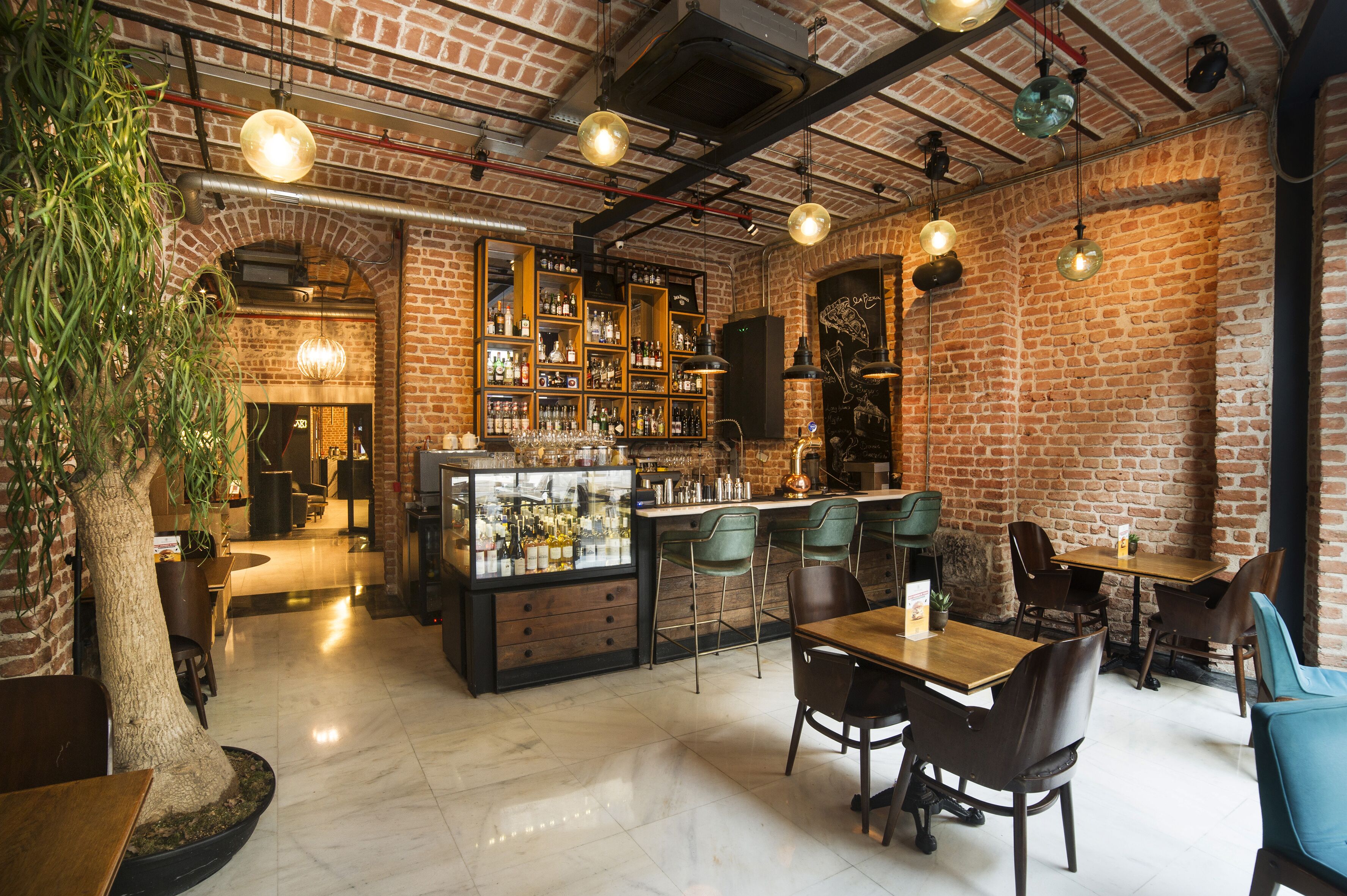 Ein Restaurant mit Backsteinwänden, Holztischen und grünen Stühlen. Es gibt eine Bar mit Regalen voller Flaschen und einer Auslage.