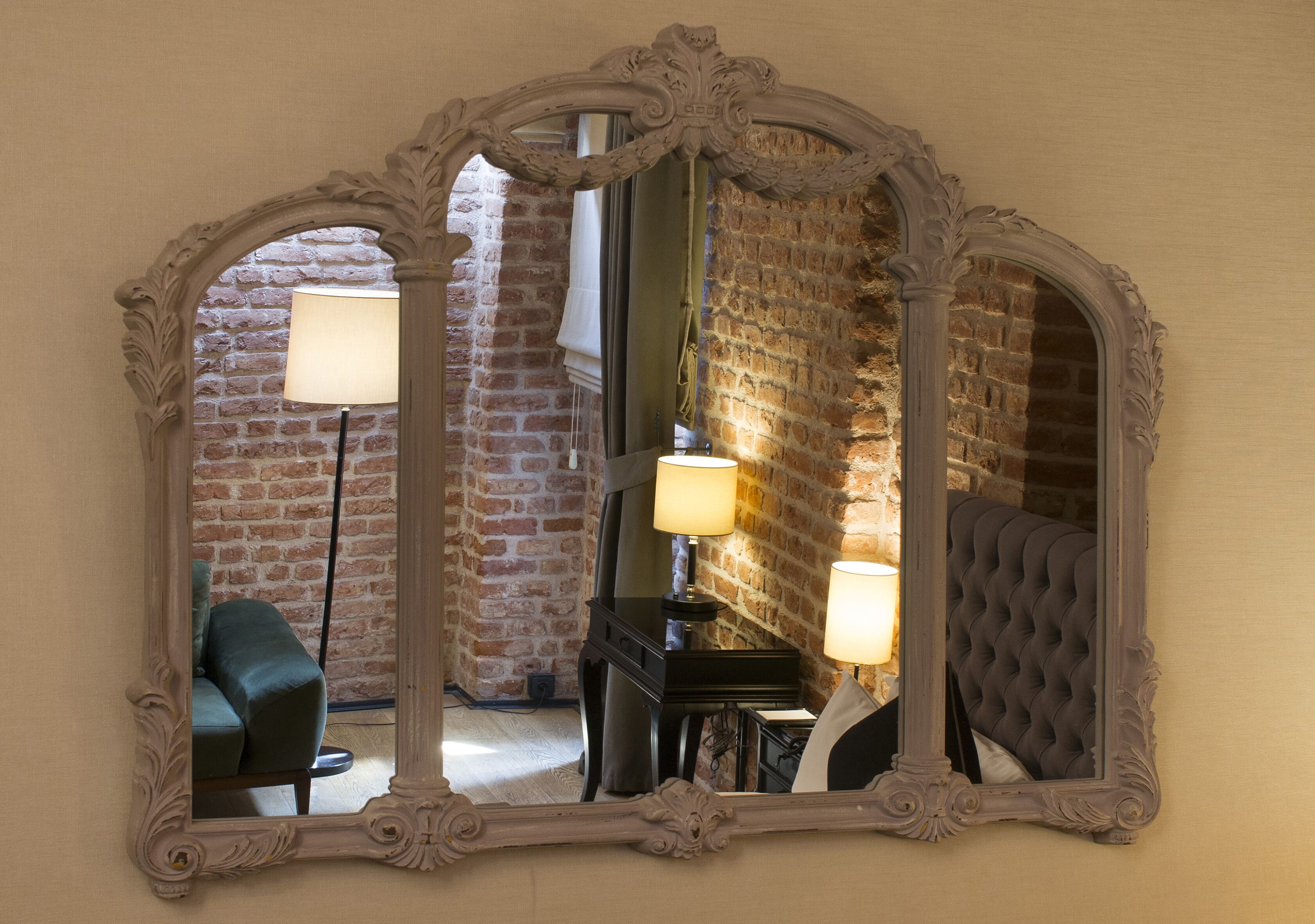 A mirror with an ornate frame reflecting a room with a piano, a lamp, and a sofa.