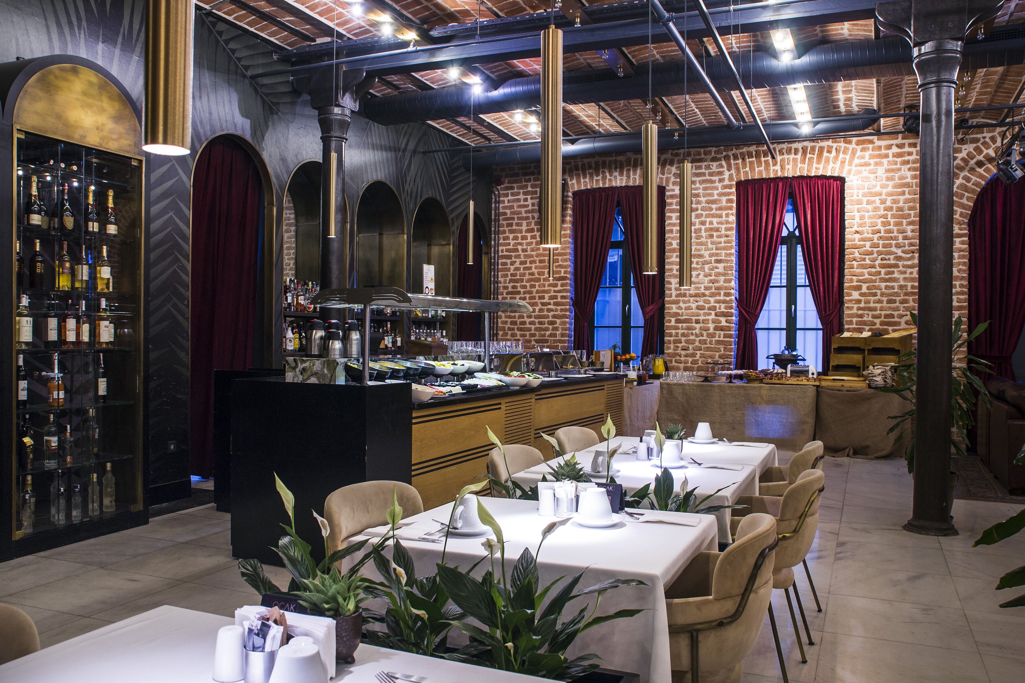 Un restaurant aux murs de briques, aux portes en arc de cercle et doté d’un bar où sont exposées de nombreuses bouteilles. Les tables sont recouvertes de nappes blanches et les chaises sont disposées autour.