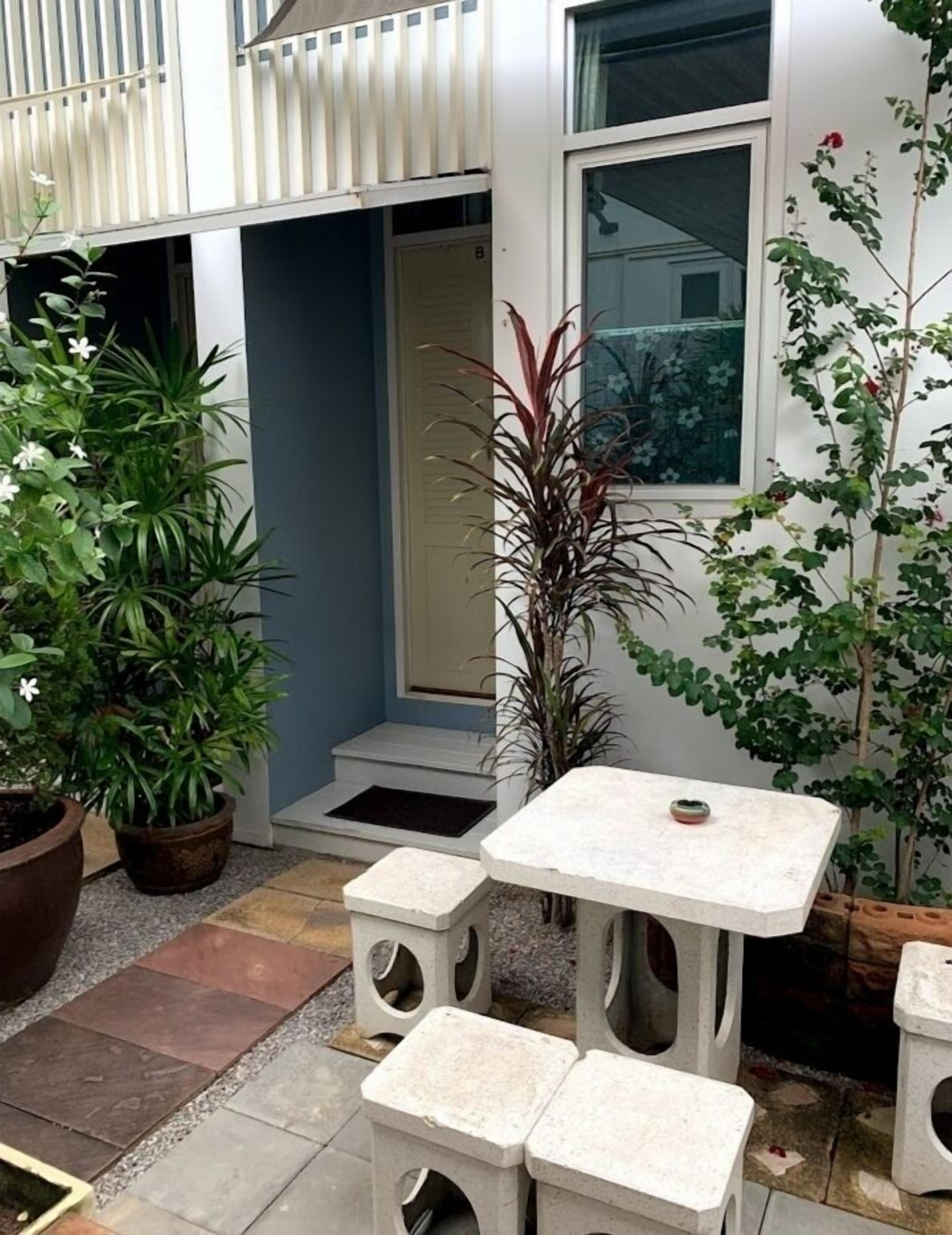 Un petit espace extérieur aménagé en patio, avec une table blanche et quatre tabourets, entouré de plantes en pot et d’un petit marchepied menant à une porte.