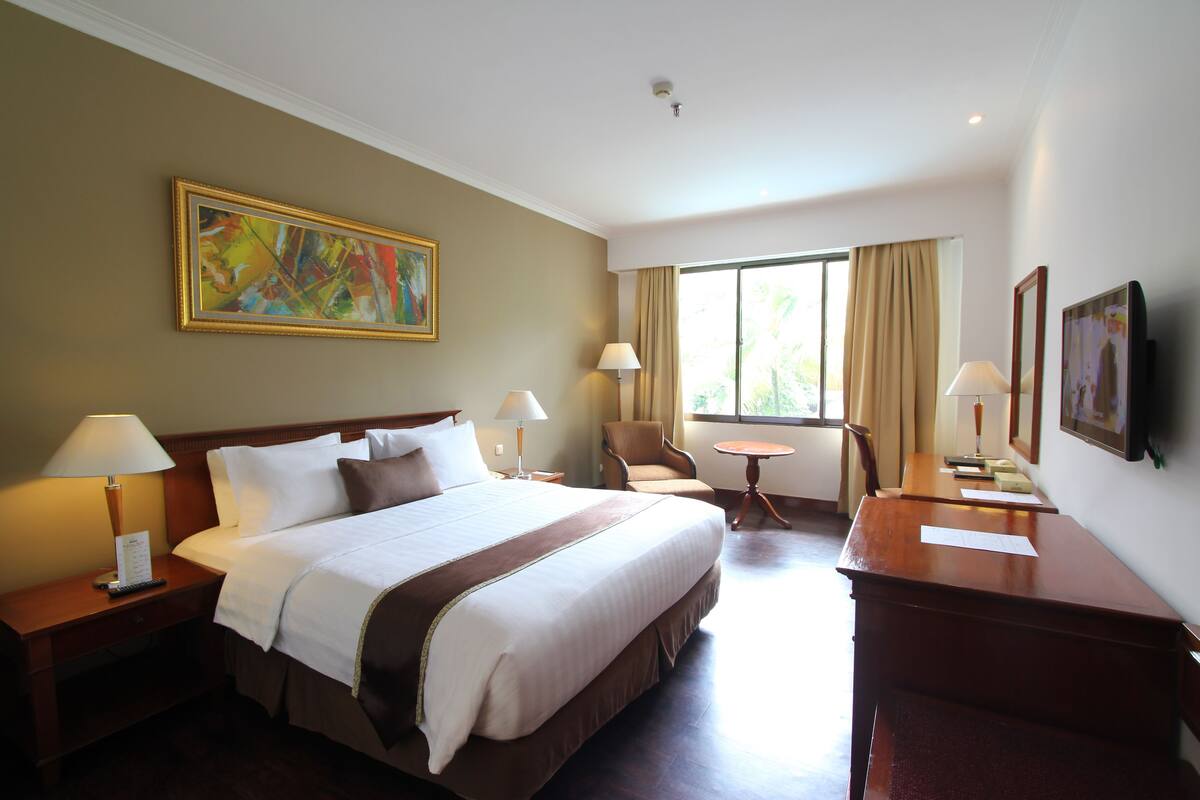 Une chambre d’hôtel avec un grand lit, deux tables de chevet avec des lampes, un bureau, une chaise, une petite table et une grande fenêtre avec des rideaux.