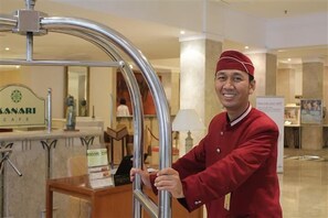 En hotellobby med en receptionsdisk, en skylt med texten ”Sanari Café” och en man i röd uniform som står vid ett metallräcke.