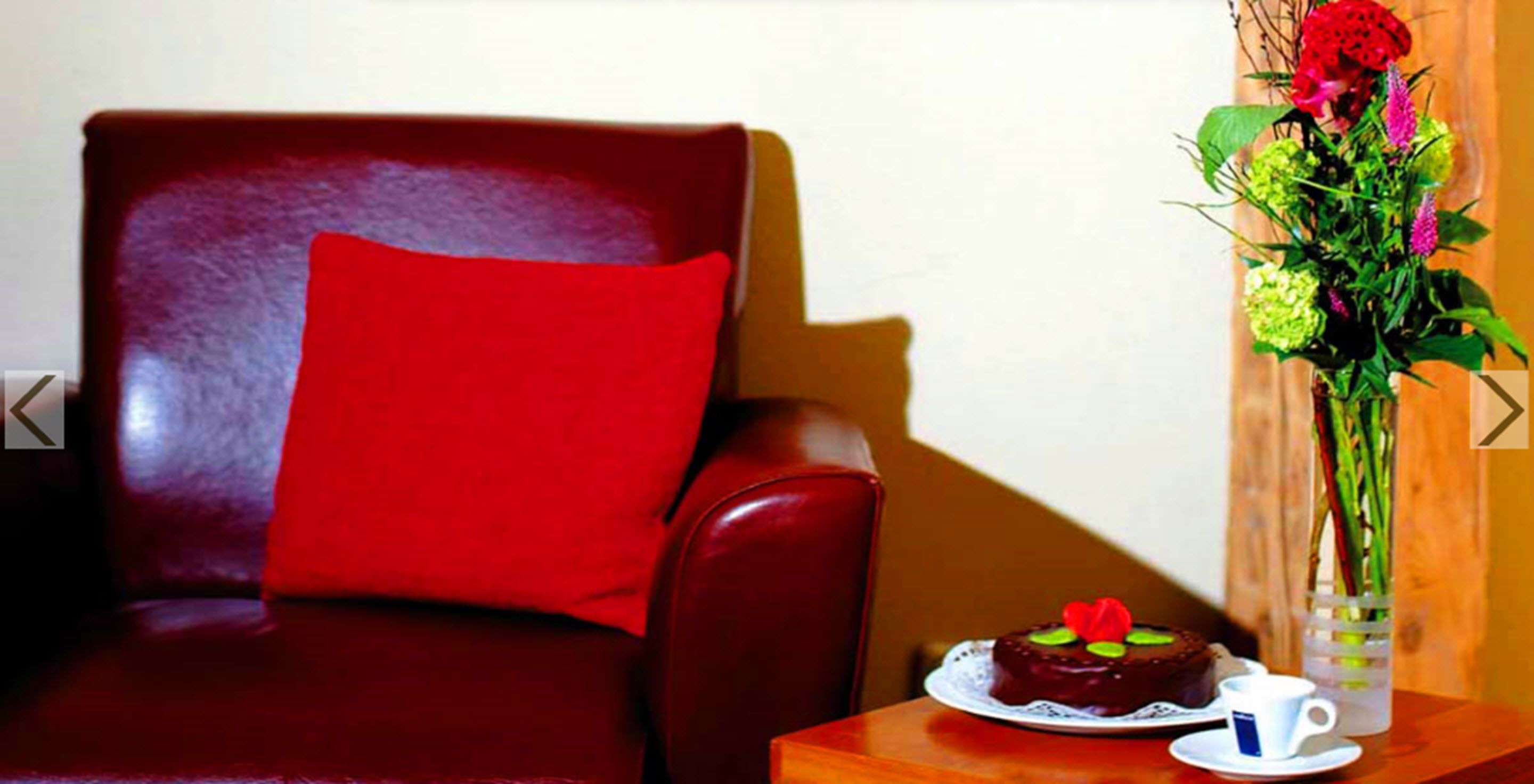 Un fauteuil en cuir rouge avec un coussin rouge, une petite table sur laquelle se trouve un gâteau au chocolat, une tasse blanche et un vase rempli de fleurs.