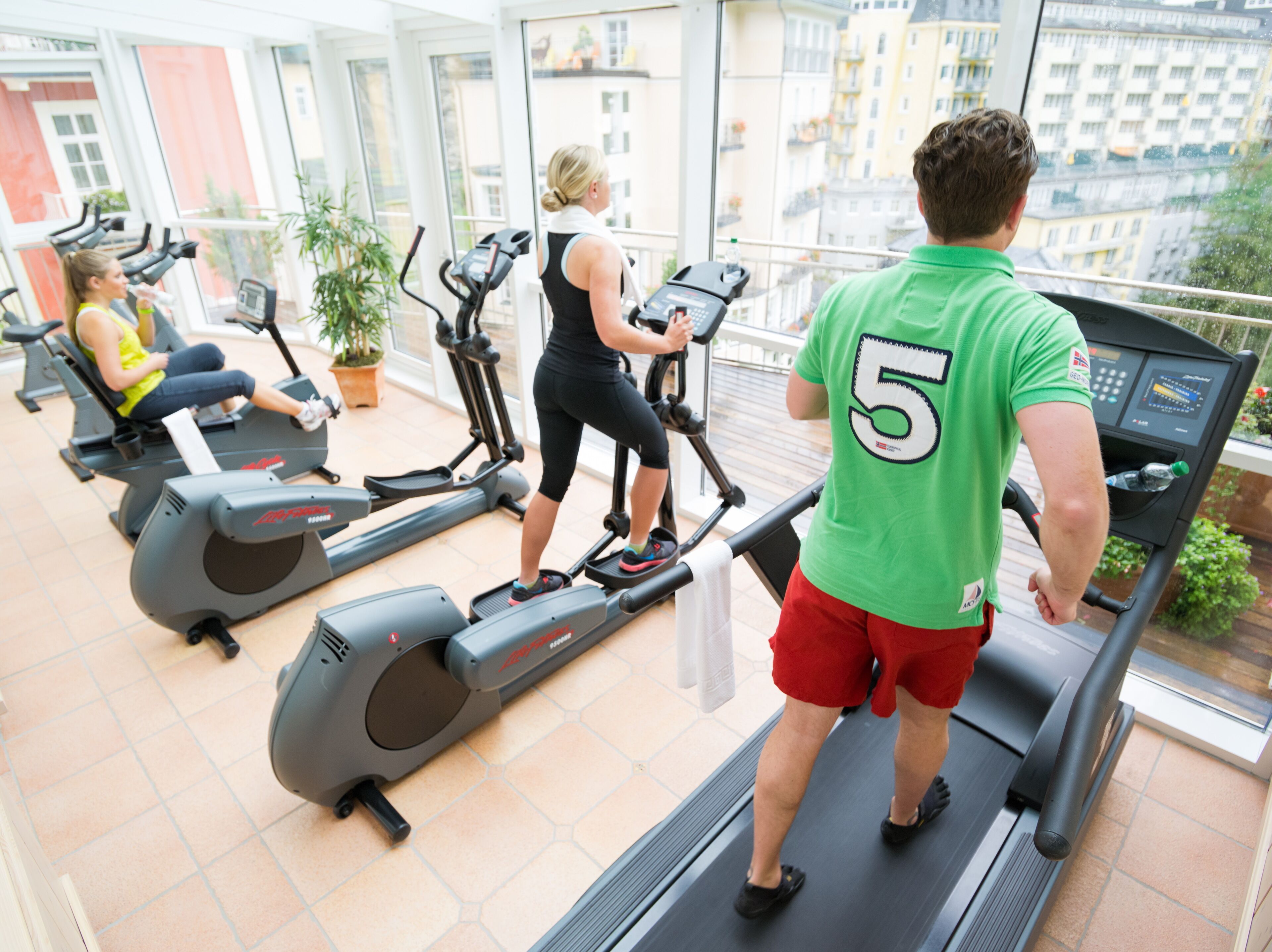 Un gimnasio con cintas de correr y bicicletas estáticas, ventanas amplias y vistas a edificios.