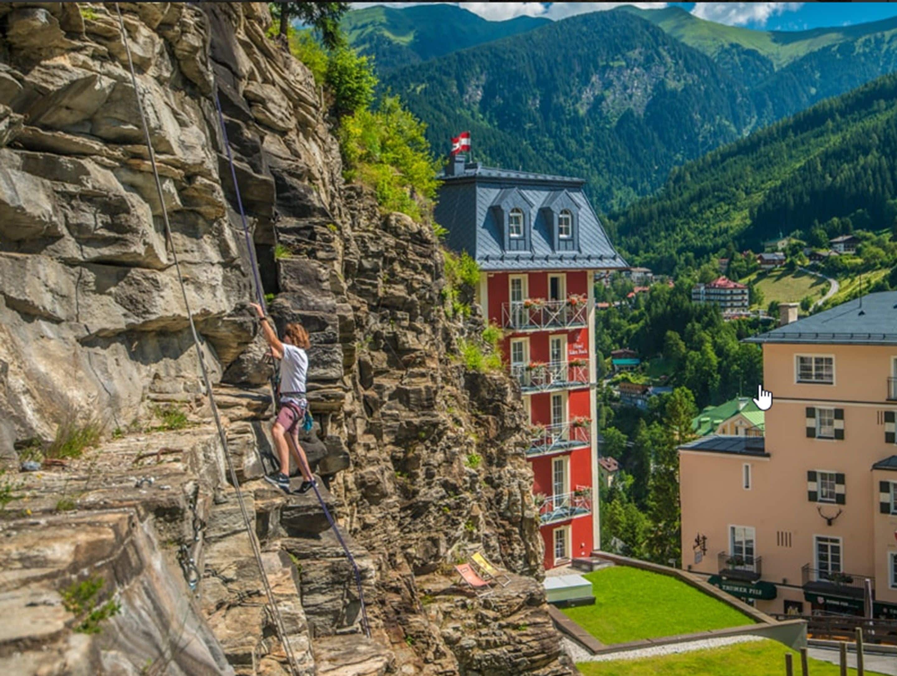 A person rock climbing on a steep rock face with a red building in the background.