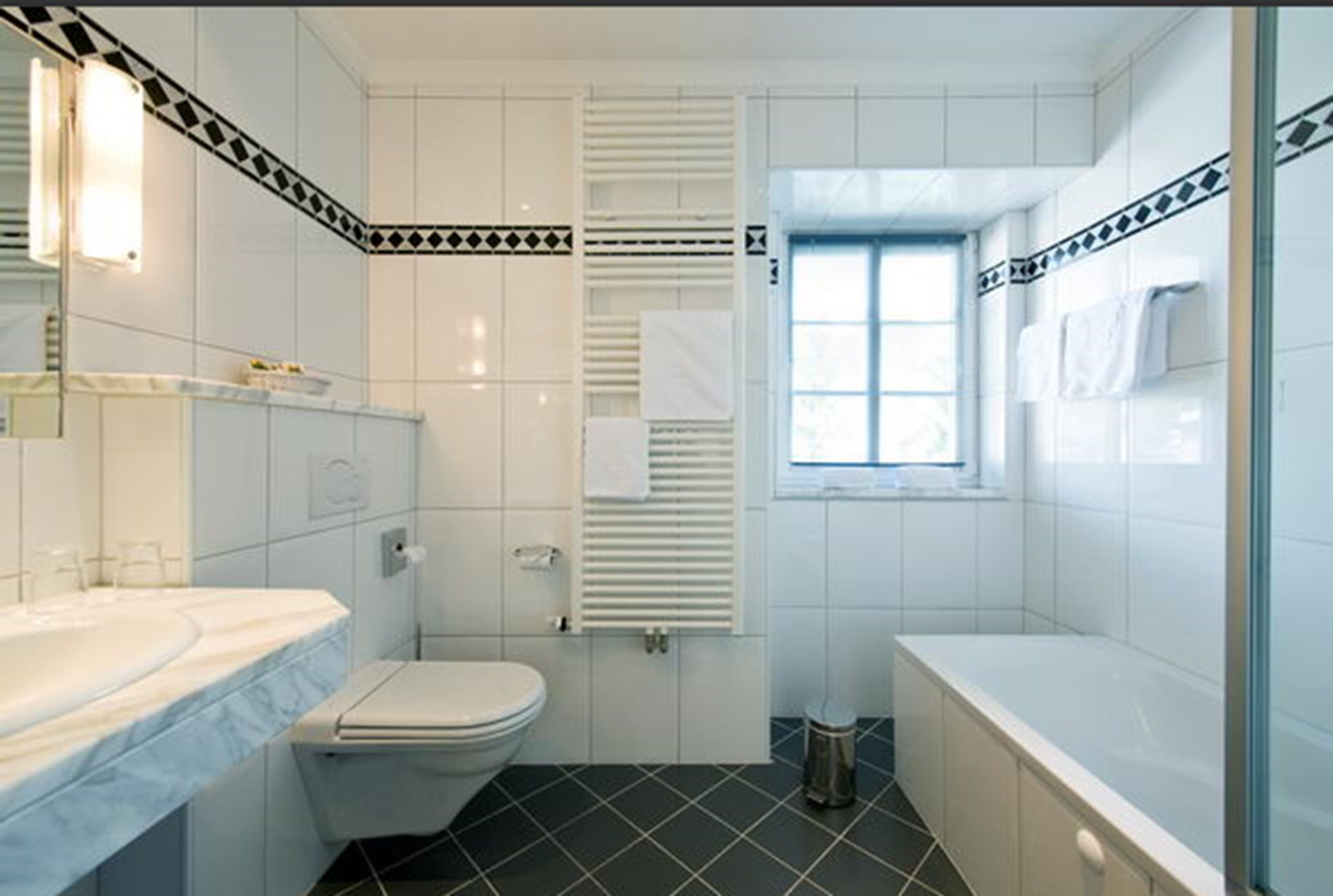 Un baño con lavamanos, inodoro y bañera blancos, suelo de baldosas y una ventana con persianas.
