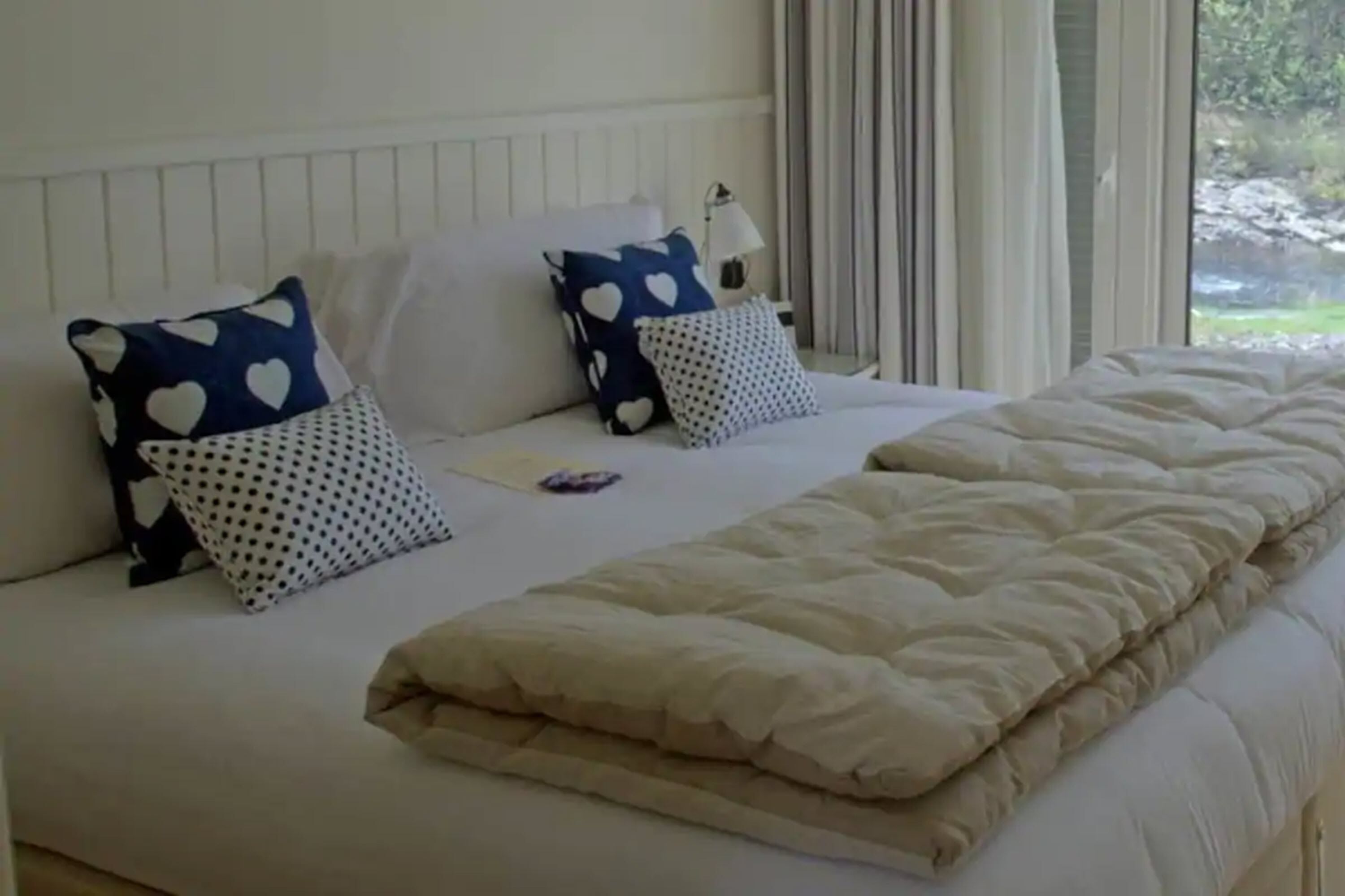 A neatly made bed with a white and beige duvet, two heart-patterned pillows, and a small bedside table with a lamp.
