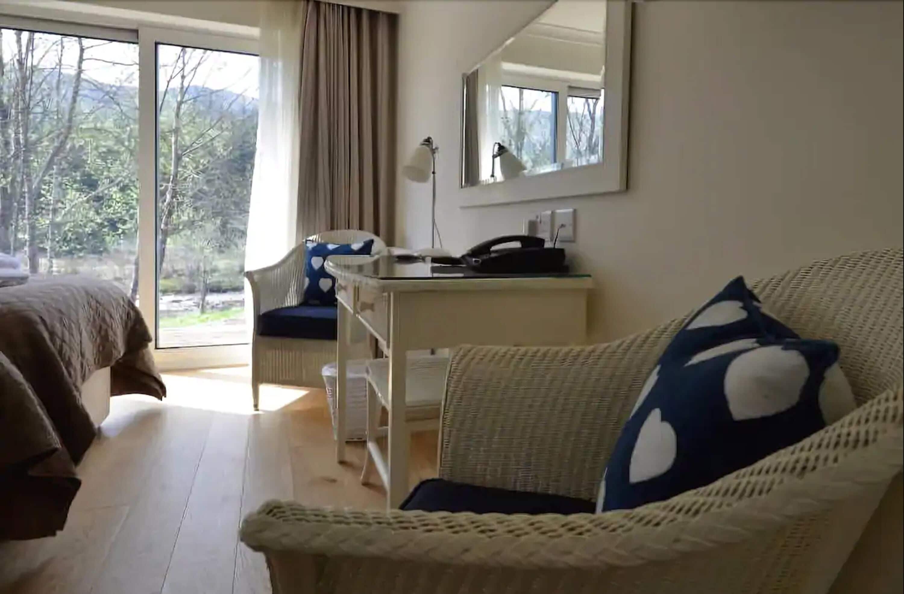 Habitación de hotel con una cama, un sofá de mimbre con un cojín estampado con corazones, un escritorio con lámpara y un ventanal con vistas a árboles y montañas.
