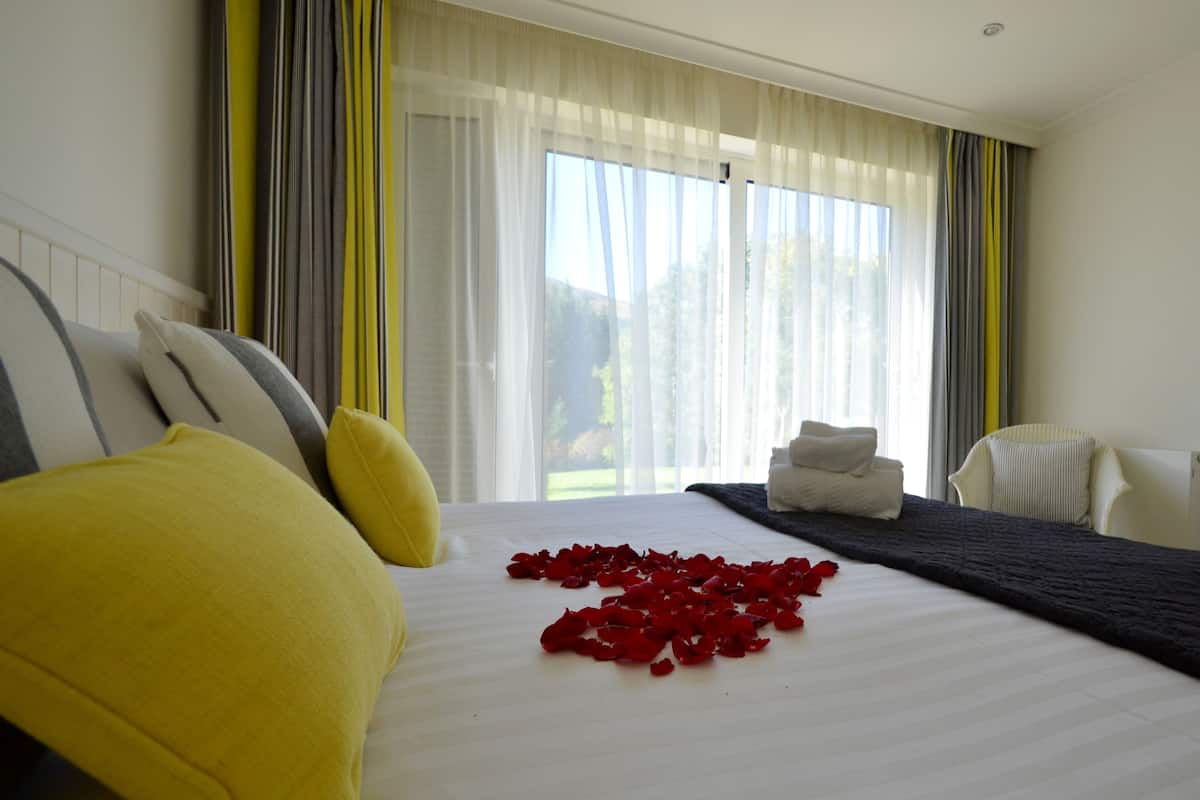 Habitación de hotel con una cama grande, dos almohadas, una mesita de noche con una toalla y vistas a zonas verdes por la ventana.