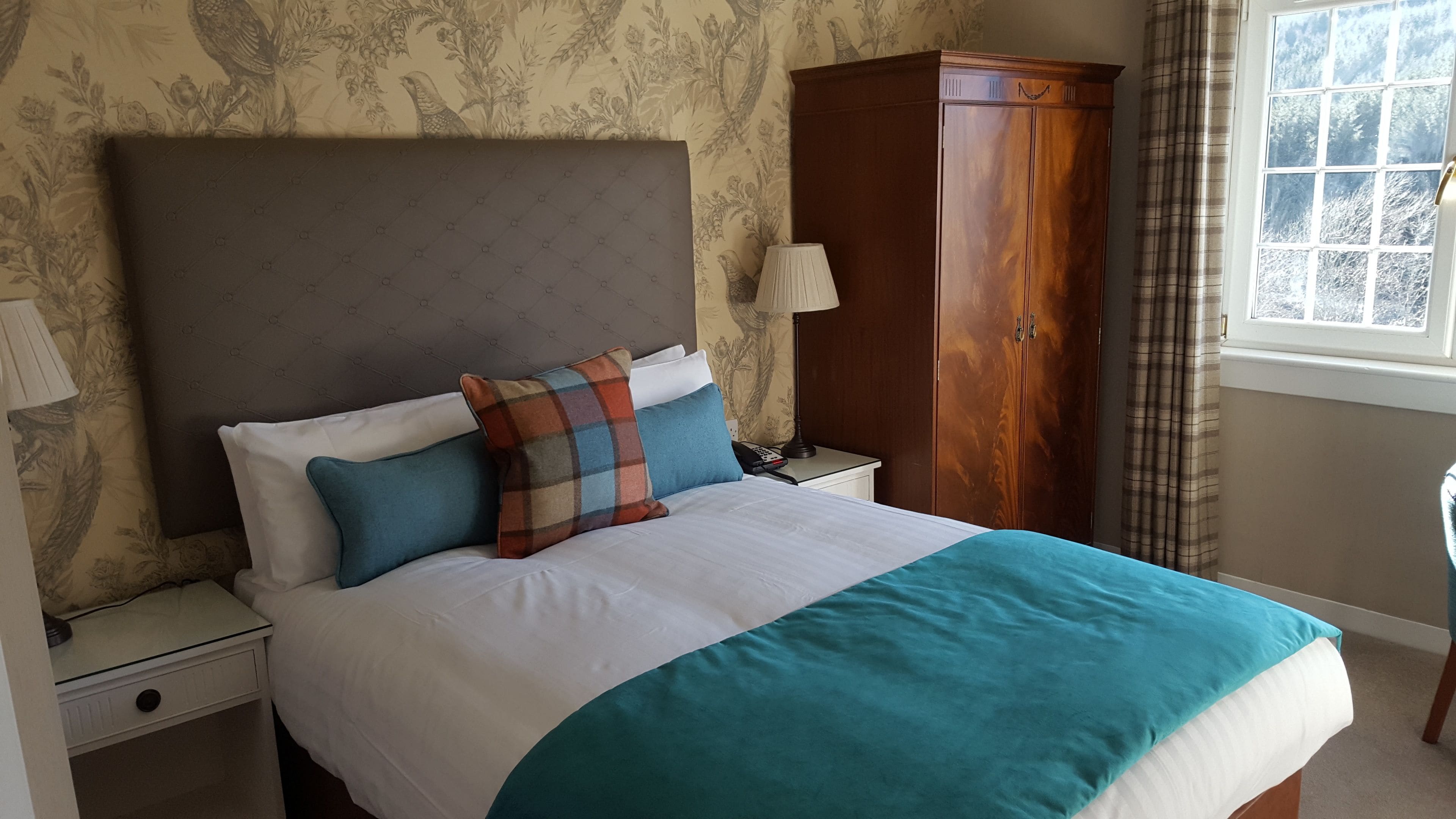 Cama bem arrumada com cabeceira estampada, guarda-roupa de madeira e mesa de cabeceira com abajur.