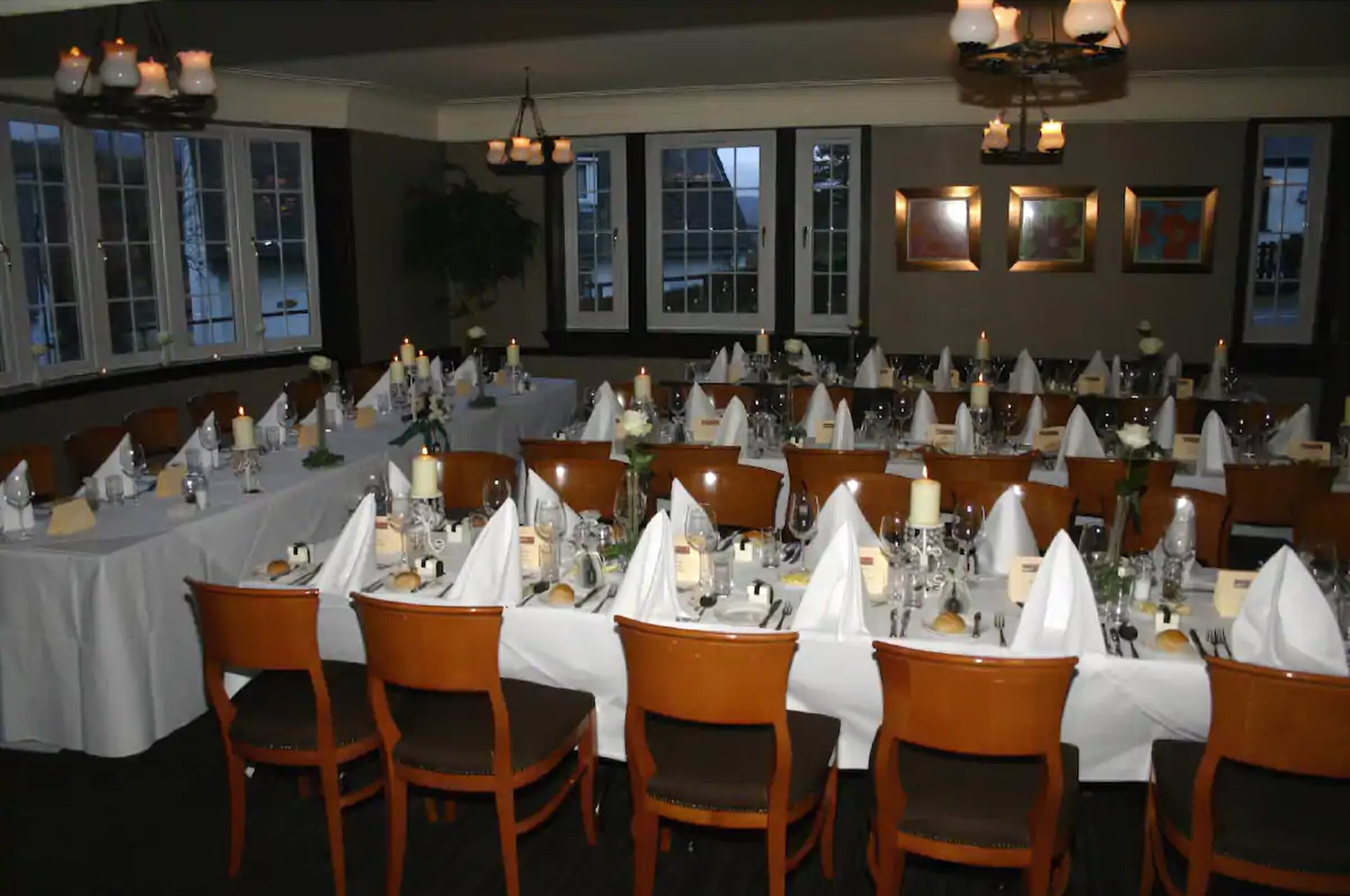 A formal dining room with multiple tables set for a meal, featuring white tablecloths, candles, and wooden chairs.