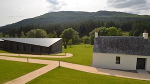 A building with a dark roof and white walls, surrounded by a well-maintained lawn and a forested hill in the background. - Bridge of Orchy Hotel (Bridge of Orchy)